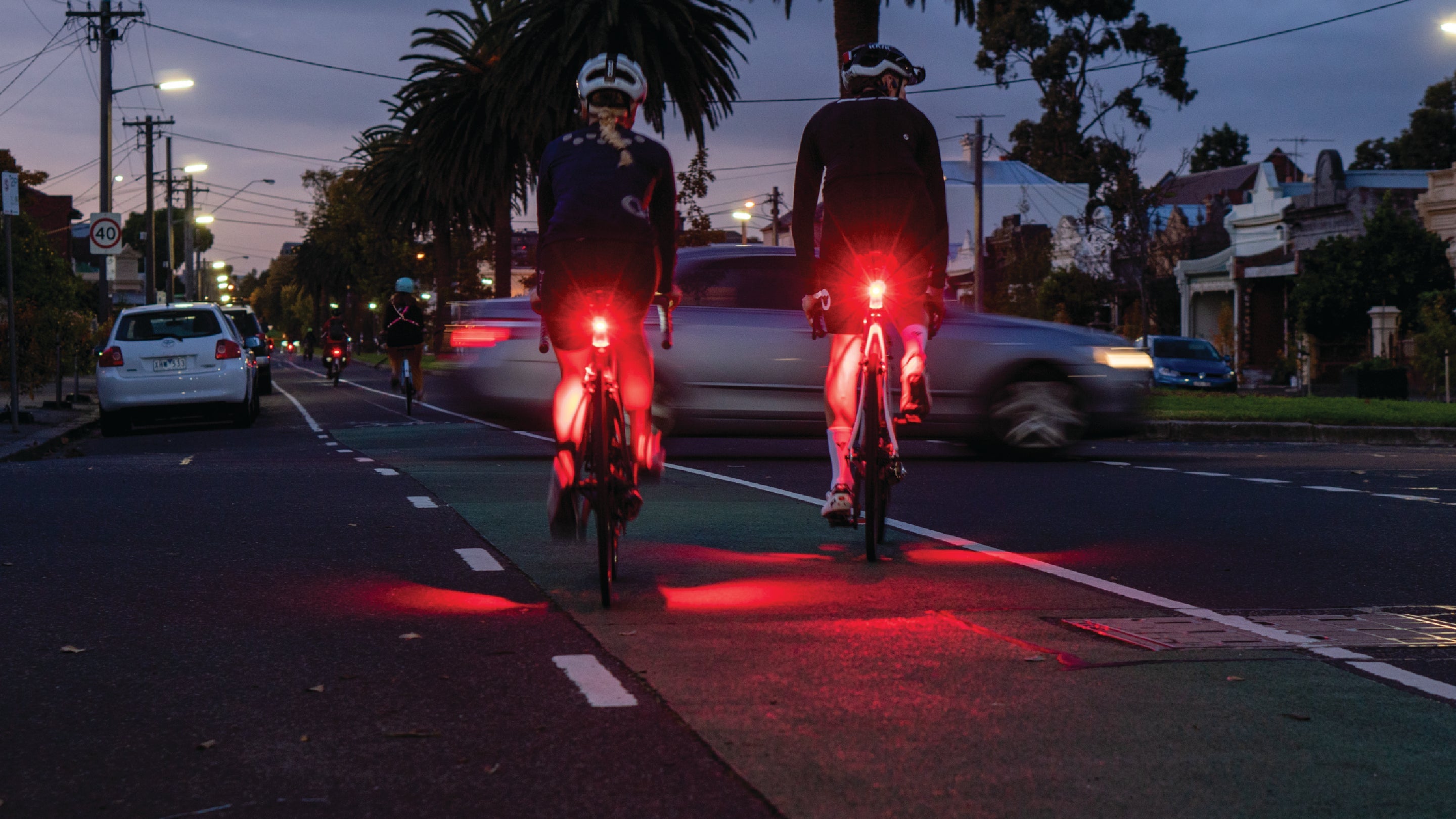 Two cyclists riding in the dark, each wearing a Flock Light emitting a bright red flashing signal for visibility. The scene captures motion with blurred streetlights in the background, highlighting the safety feature of the Flock Light during nighttime cycling. The cyclists wear casual dark clothing and helmets. Available at the RMIT Store.